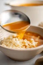 Large stainless steel ladle pouring broth into a wide soup bowl filled with cooked pastina pasta.