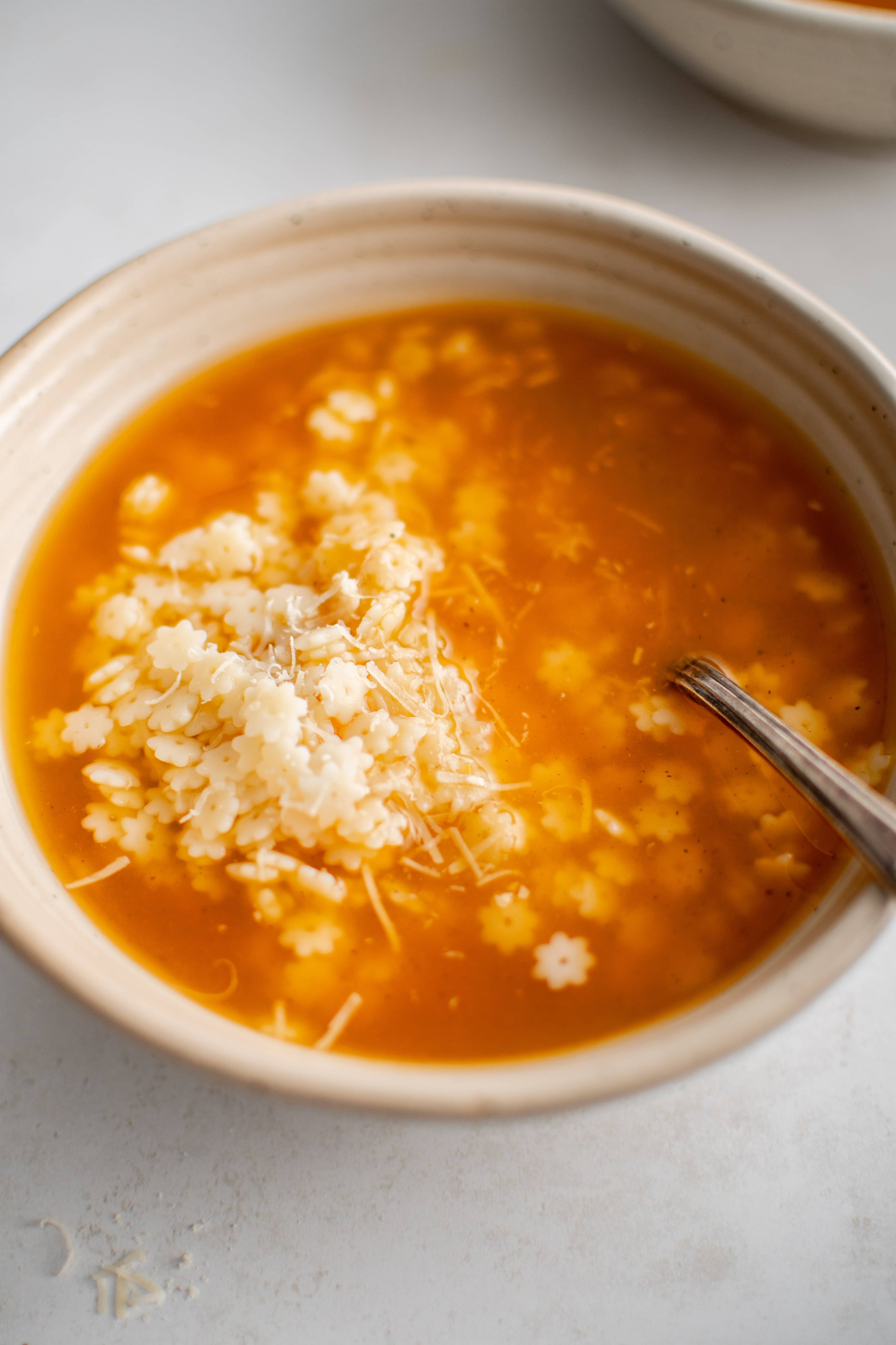 Beige colored soup bowl filled with pastina in a rich and comforting orange-colored vegetable chicken broth and garnished with freshly grated parmesan cheese.
