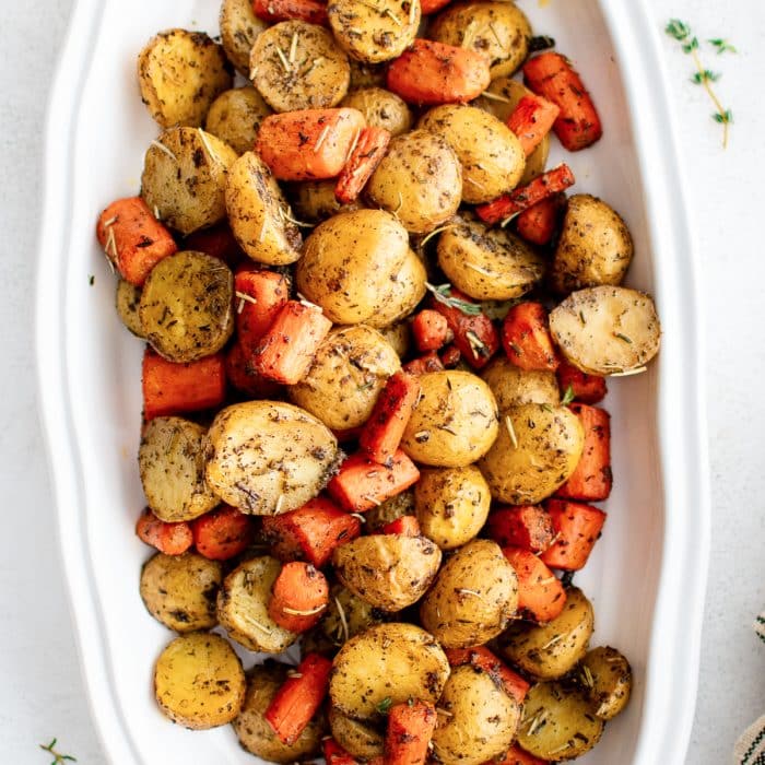Roasted carrots and baby potatoes served family-style in a large white platter with flecks of seasoning and fresh thyme sprigs.
