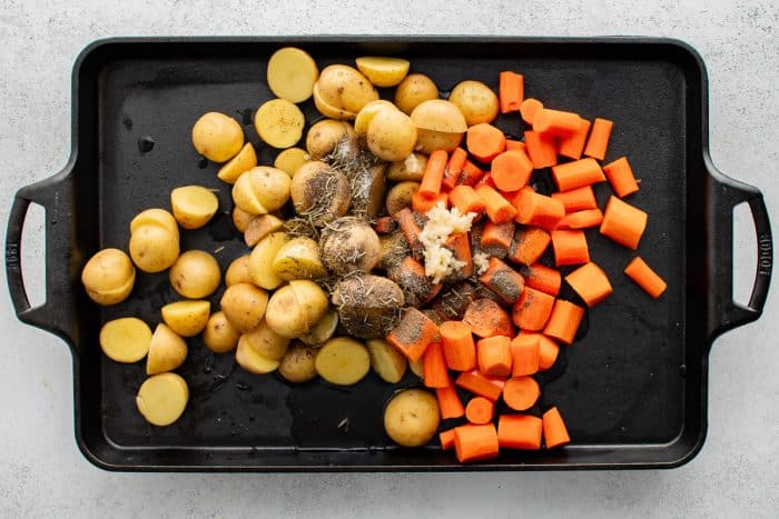 Uncooked baby potatoes, carrots, garlic, and seasonings on a black baking pan.