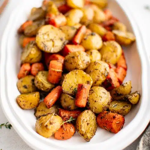 Large white oval serving platter filled with golden roasted potatoes and carrots coated in dried rosemary, thyme, salt, black pepper, olive oil, and fresh toasted garlic.