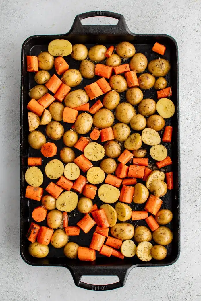Large Lodge cast iron baking sheet pan covered with a single layer of halved baby potatoes and carrots coated in olive oil, minced garlic, dried rosemary and dried thyme, salt, and black pepper.