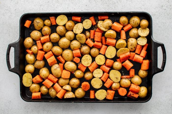 Large Lodge cast iron baking sheet pan covered with a single layer of halved baby potatoes and carrots coated in olive oil, minced garlic, dried rosemary and dried thyme, salt, and black pepper.