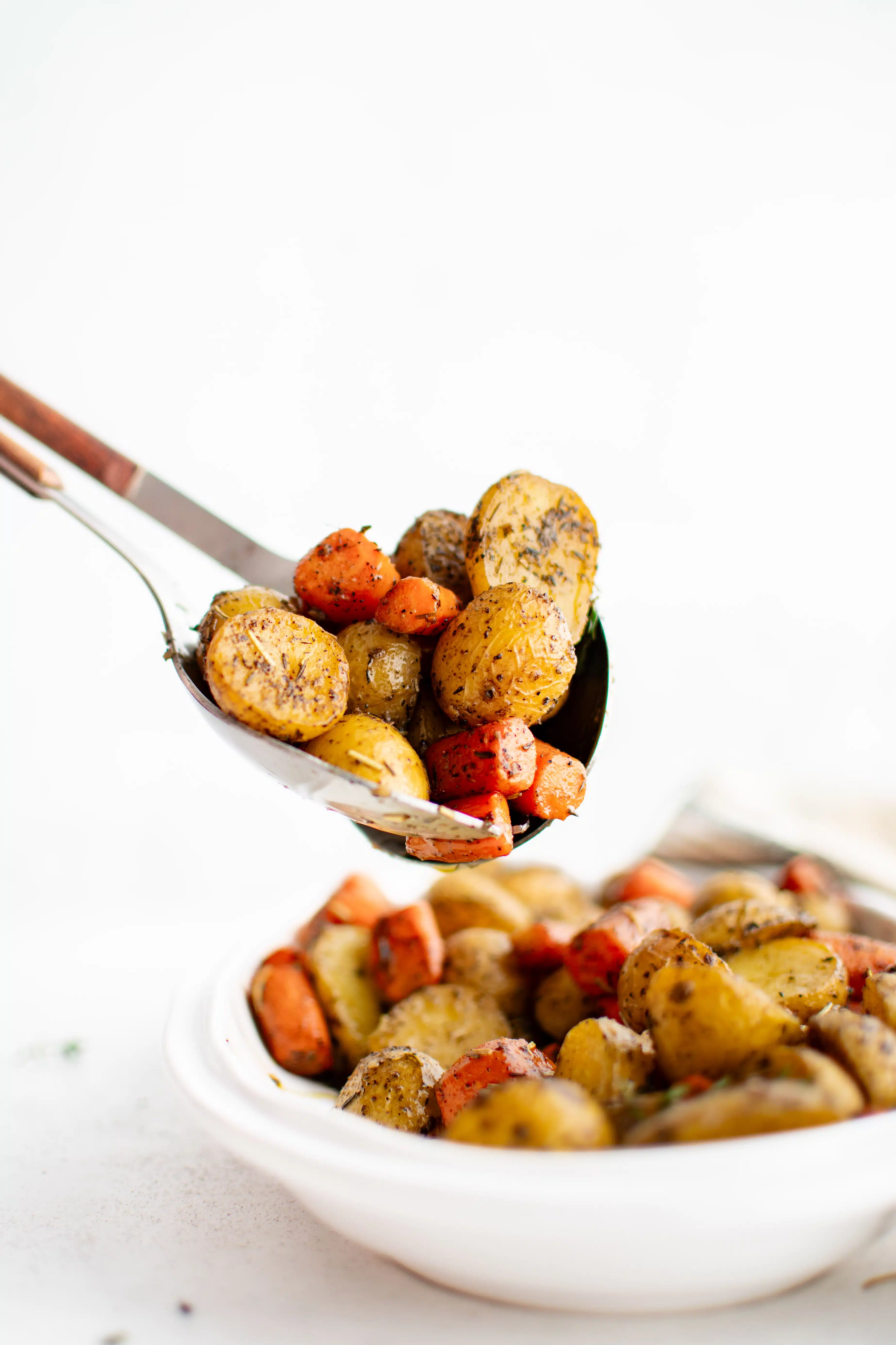 Large serving spoon with a scoop of roasted potatoes and carrots taken from a large white serving dish filled with the remaining roasted veggies.