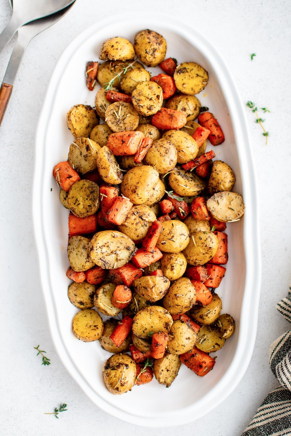 Roasted carrots and baby potatoes served family-style in a large white platter with flecks of seasoning and fresh thyme sprigs.