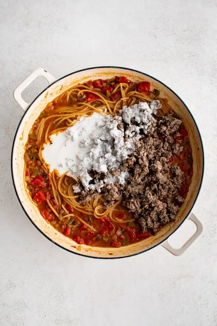 Cooked ground beef and crema added to a large pot filled with cooked spaghetti noodles combined with a mixture of onions, Rotel, and seasonings and spices.