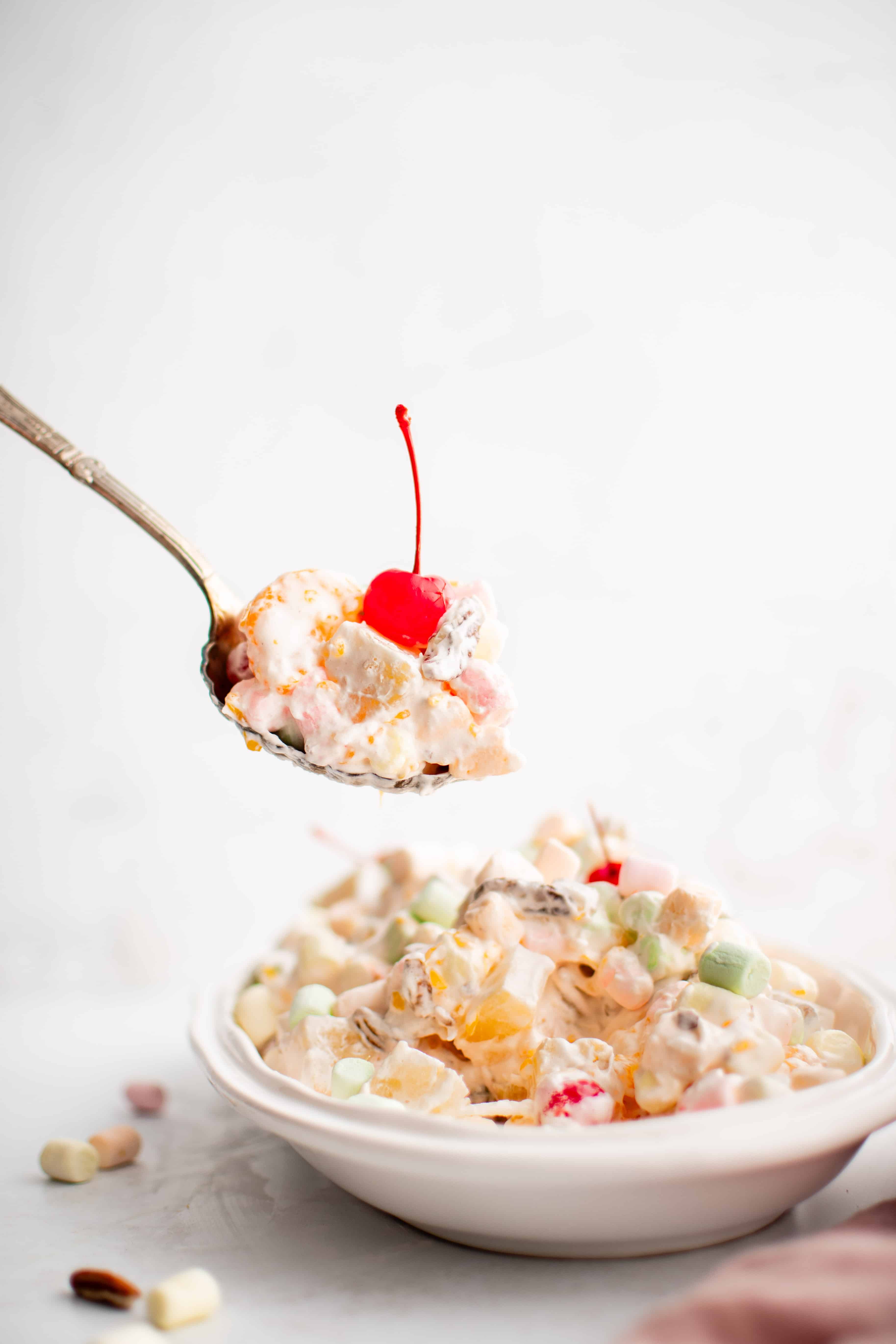 Large spoonful of ambrosia salad scooped from a large oval platter filled with mini marshmallows, pineapple, mandarin oranges, grapes, maraschino cherries, pecans, and shredded coconut in a creamy whipped topping.