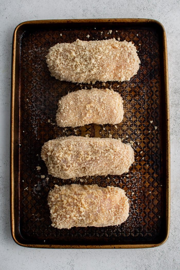 Four breadcrumb coated rolled-up ham and swiss cheese stuffed chicken breasts on a large baking sheet.