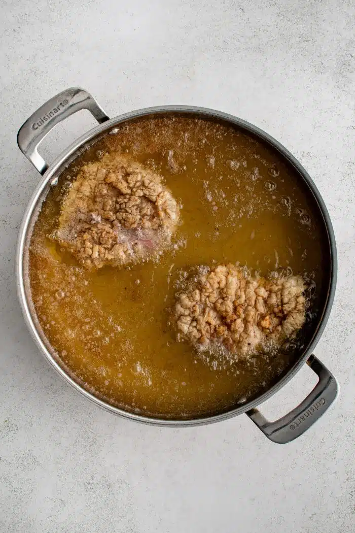 Two breaded chicken fried steaks frying in a large pan filled with hot oil.