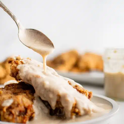 Spoon drizzling homemade creamy white gravy over two pieces of chicken fried steak on a white dinner plate.