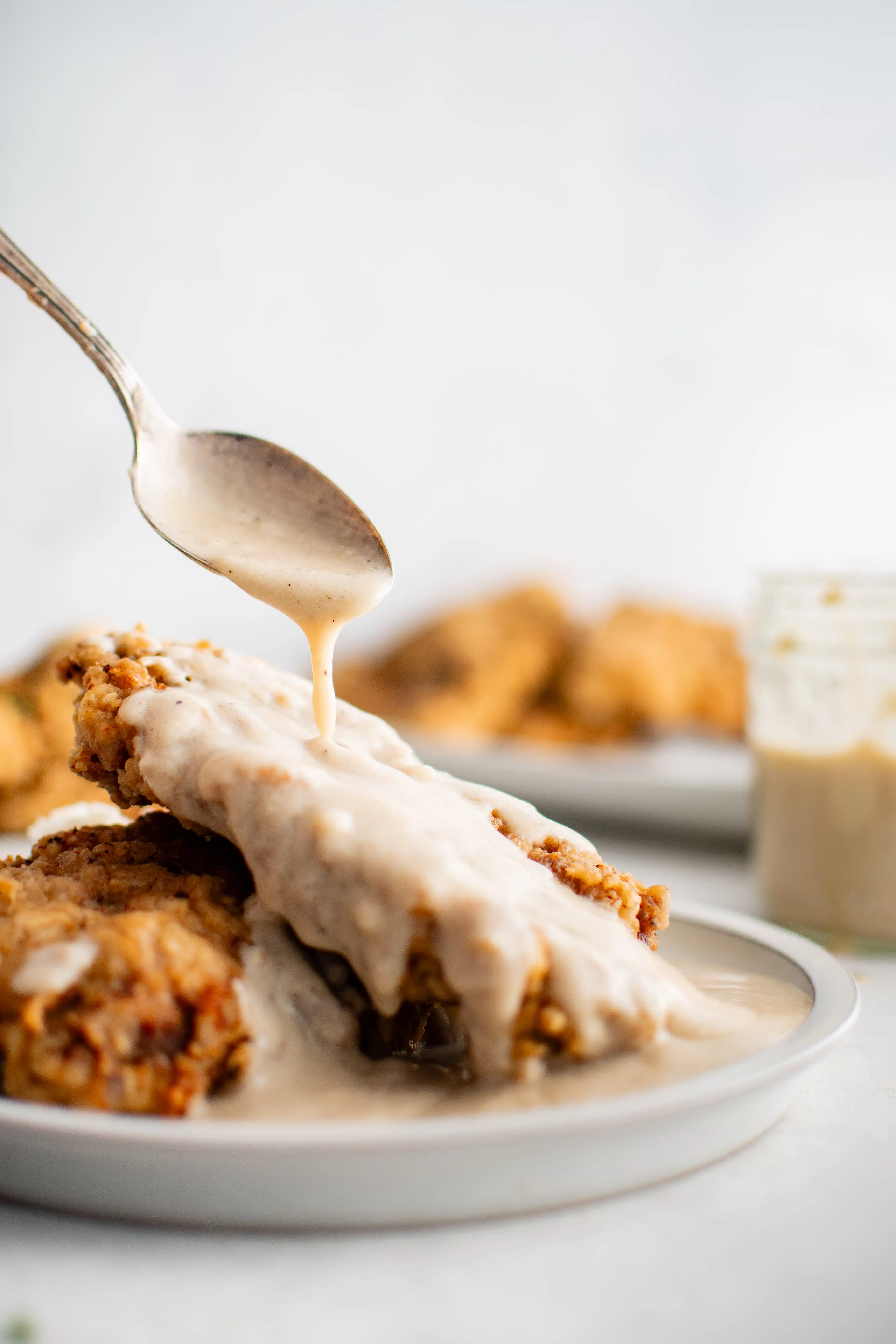 Spoon drizzling homemade creamy white gravy over two pieces of chicken fried steak on a white dinner plate.