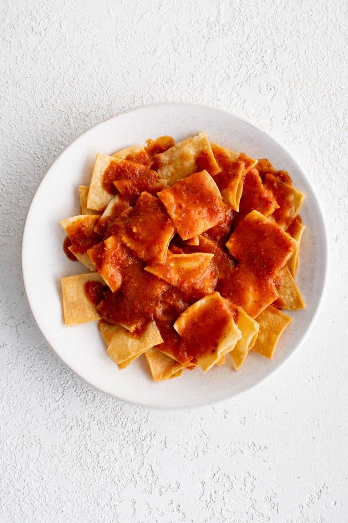 White plate filled with fried corn tortilla chips smothered in red chilaquiles sauce.