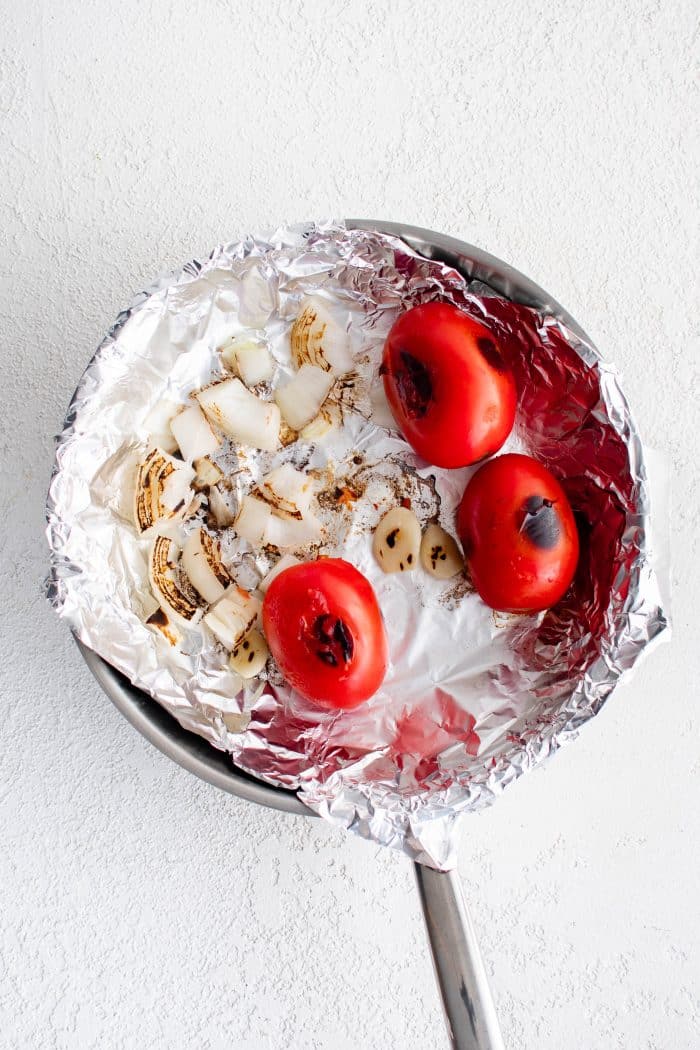 Skillet lined with aluminum foil with charred onion and three charred Roma tomatoes.