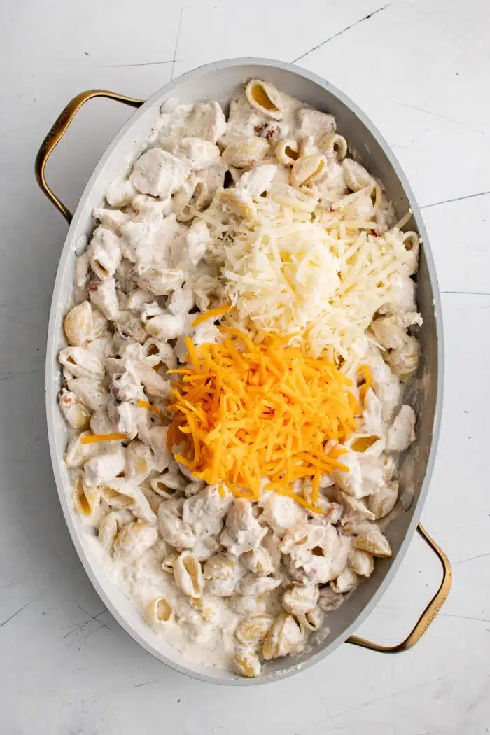 Cream cheese and ranch coated chicken, cooked pasta shells, and bacon in a large non-stick casserole dish and topped with a small pile of shredded mozzarella cheese and cheddar cheese.