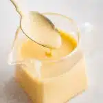 Small glass jar filled with homemade hollandaise sauce with a small spoon filled with hollandaise sauce hovering above.