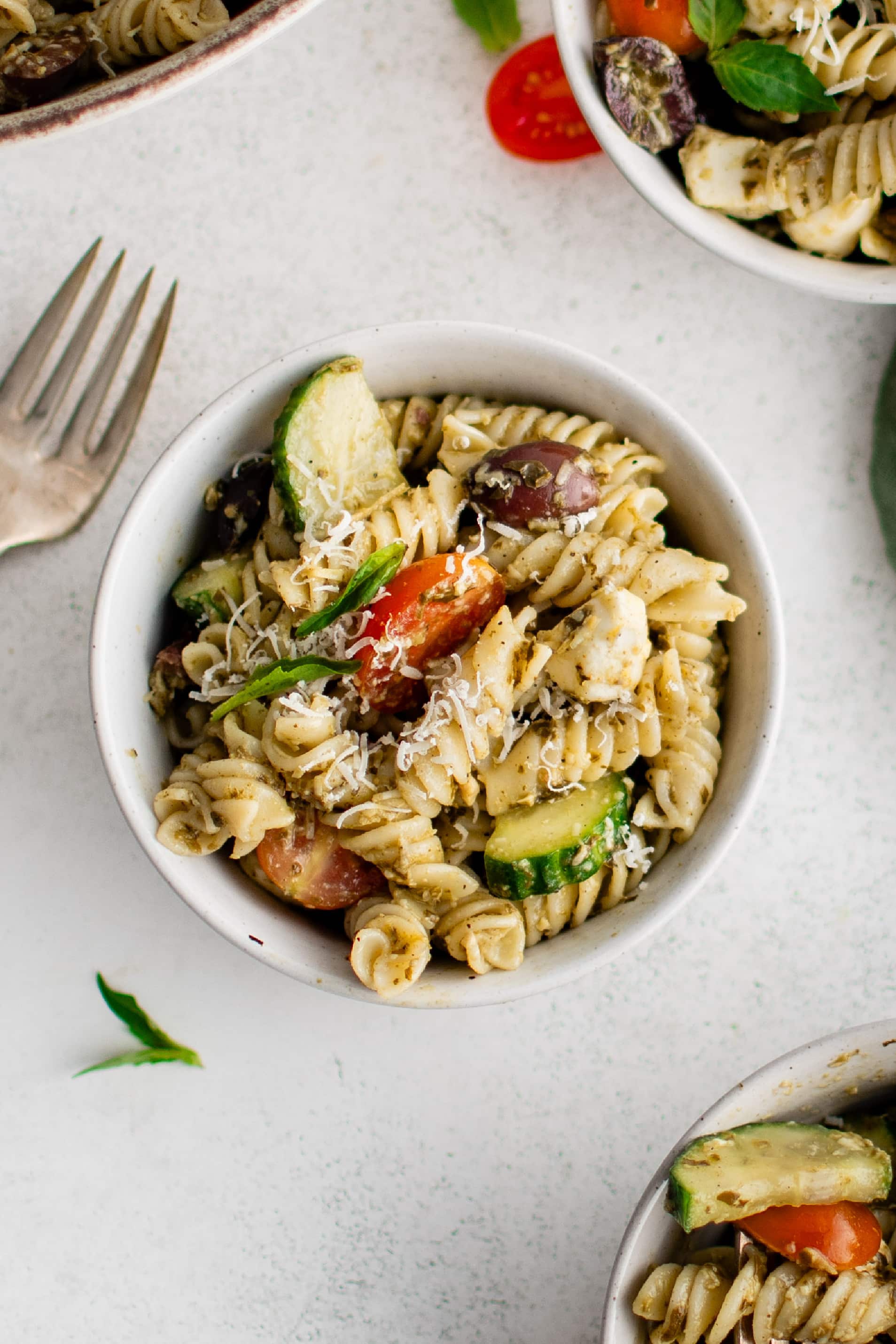 Small white bowl filled with an individual serving of cold pesto pasta salad made with rotini pasta, tomatoes, cucumber, mozzarella pearls, and kalamata olives.