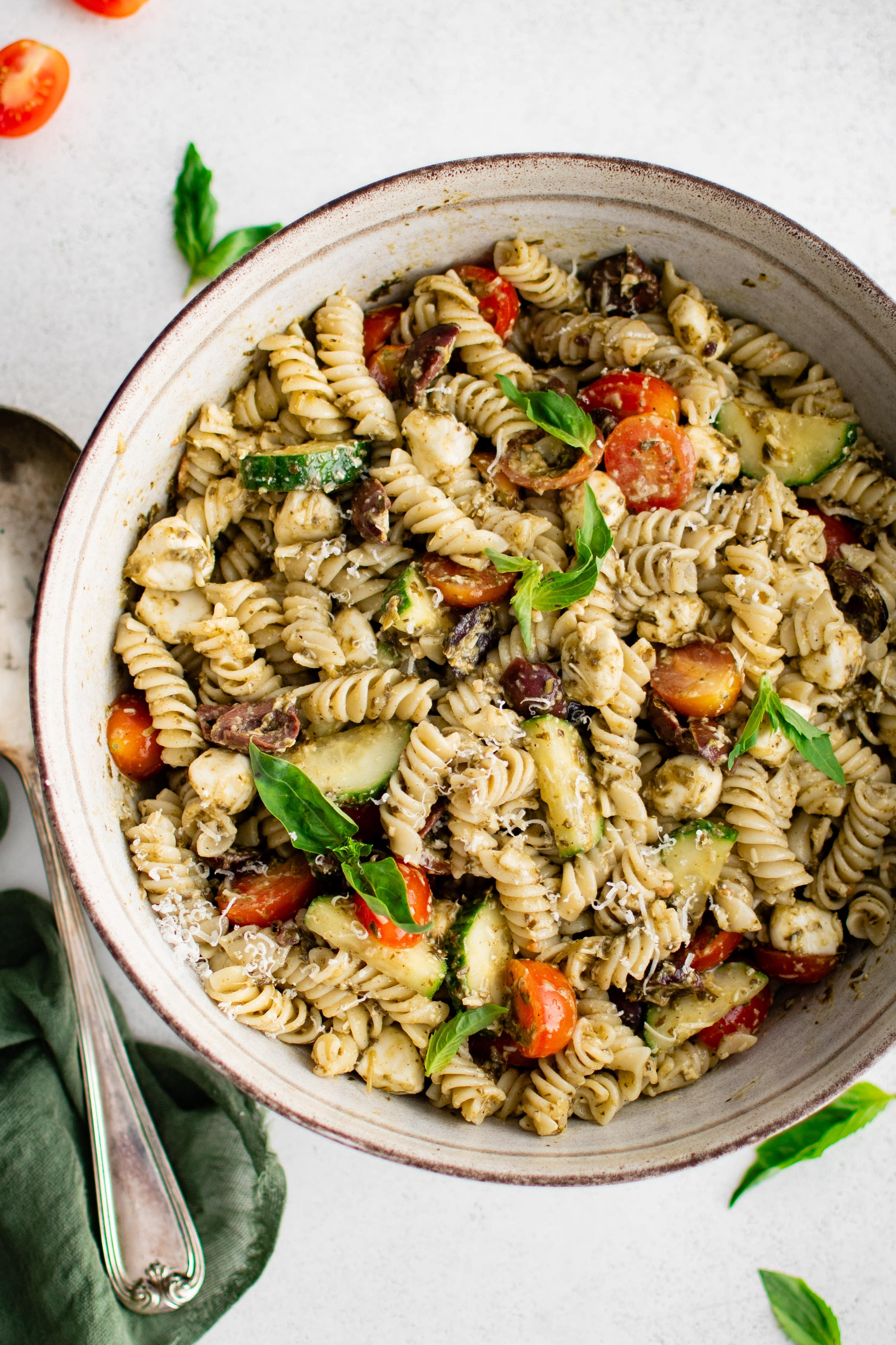 Large serving bowl filled with cold pesto pasta salad made with rotini pasta, tomatoes, cucumber, mozzarella pearls, and kalamata olives.