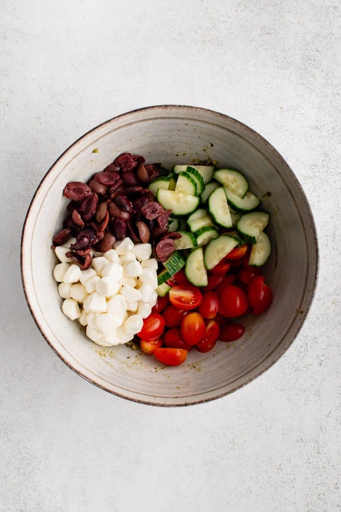 Large mixing bowl filled with halved kalamata olives, halved and sliced cucumbers, halved cherry tomatoes, and mozzarella cheese pearls.