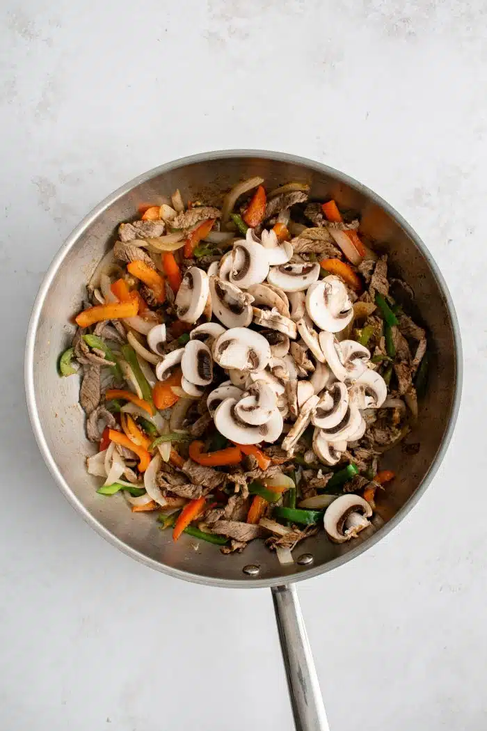 Sliced mushrooms, hot sauce, and Worcestershire sauce added to a large skillet filled with sauteed sliced onion, red and green bell peppers, and sirloin steak.