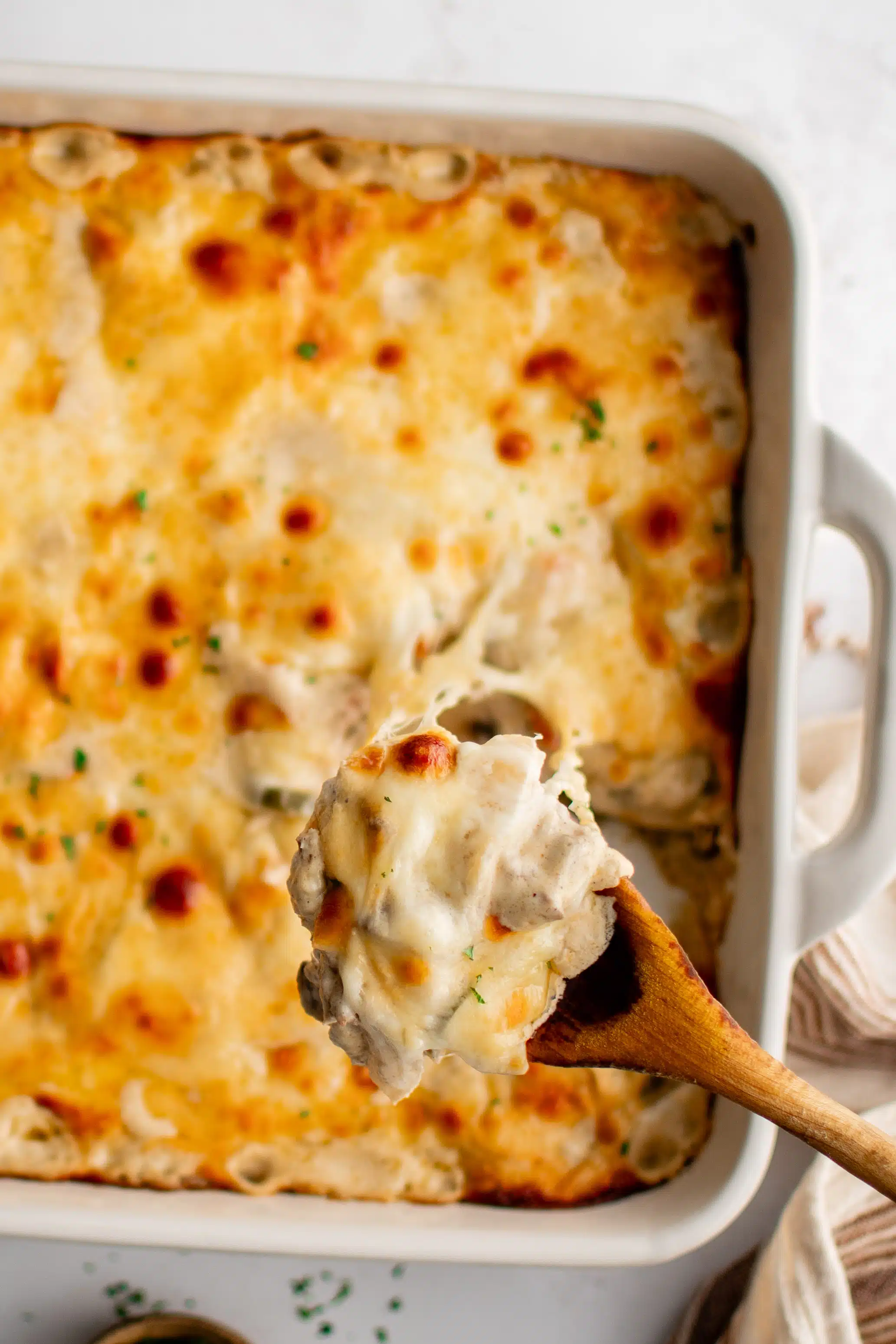 Large wooden spoon filled with Philly cheesesteak casserole hovering above a large casserole dish filled with philly cheesesteak casserole covered in gooey melted cheese.