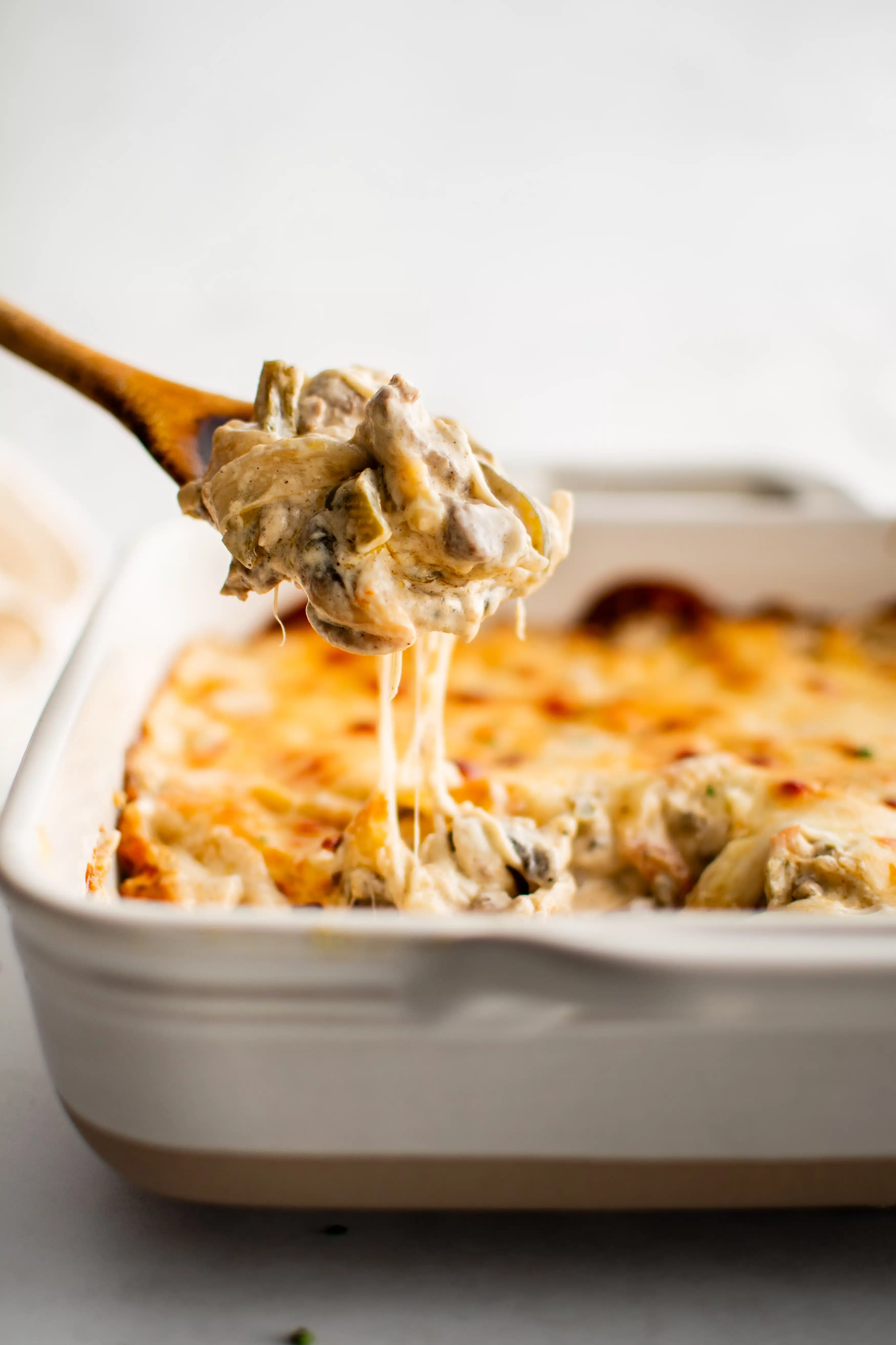 Large wooden spoon filled with Philly cheesesteak casserole hovering above a large casserole dish filled with philly cheesesteak casserole covered in gooey melted cheese.