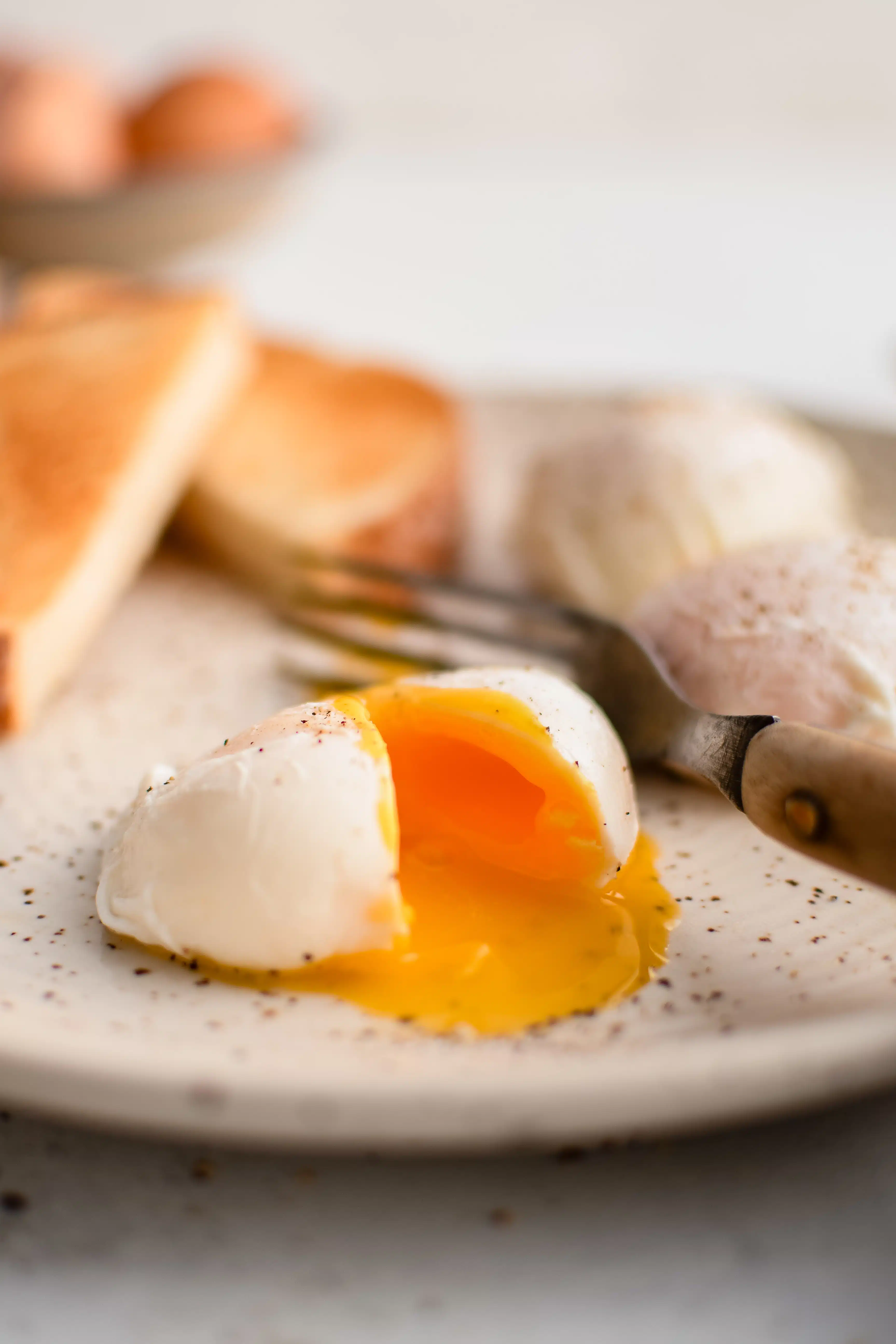 Poached egg sliced open with a runny yellow yolk on a white plate with sliced toast and two more poached eggs in the background.