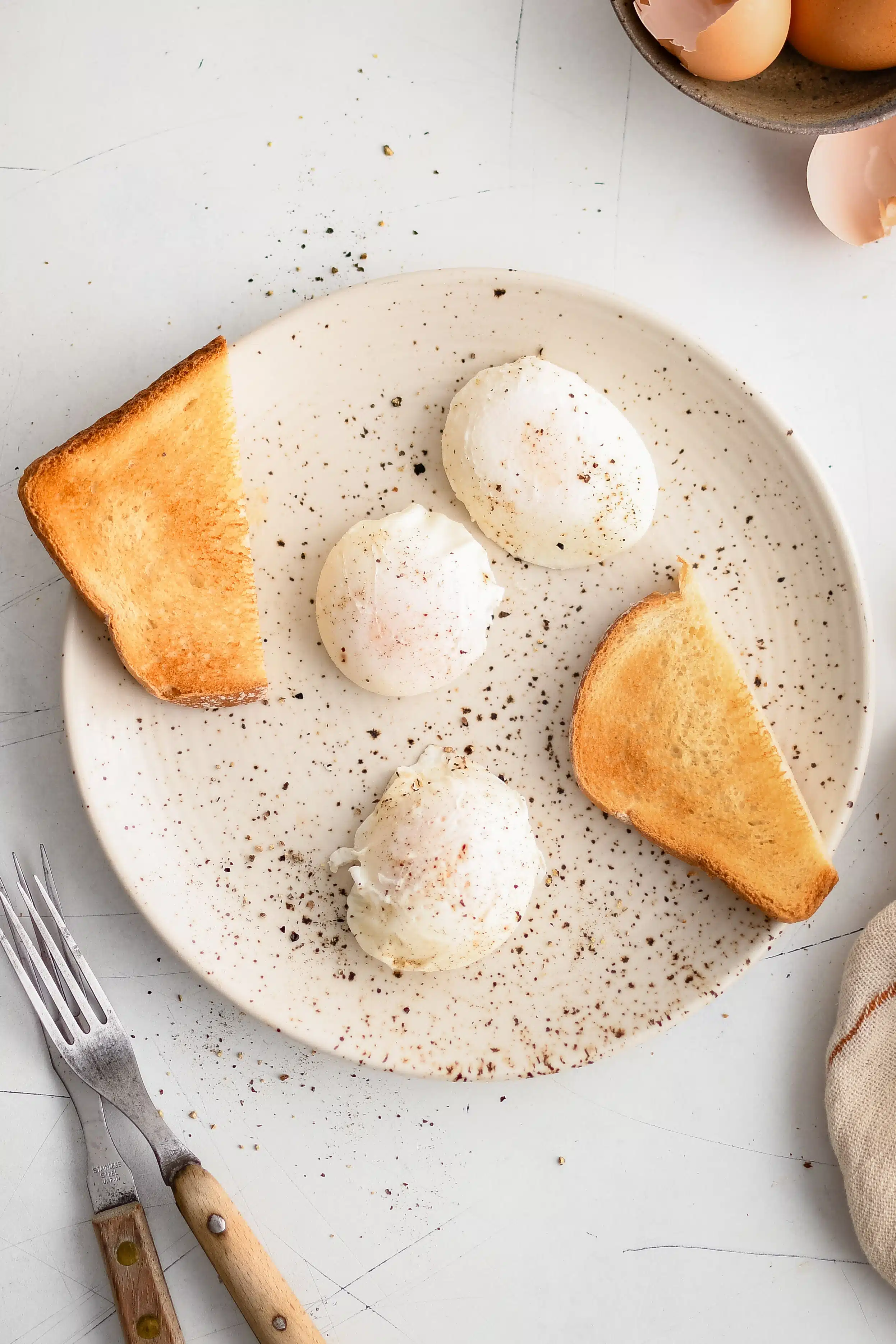 White plate with three poached eggs seasoned with black pepper and a piece of toasted bread sliced in half.