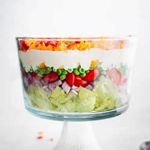 Beautifully layered traditional 7 layer salad starting with chopped iceberg lettuce on the bottom, followed by chopped red onion, sliced cherry tomatoes, green peas, chopped hard boiled eggs, creamy mayonnaise dressing, shredded cheddar cheese, and bacon bits on the top.