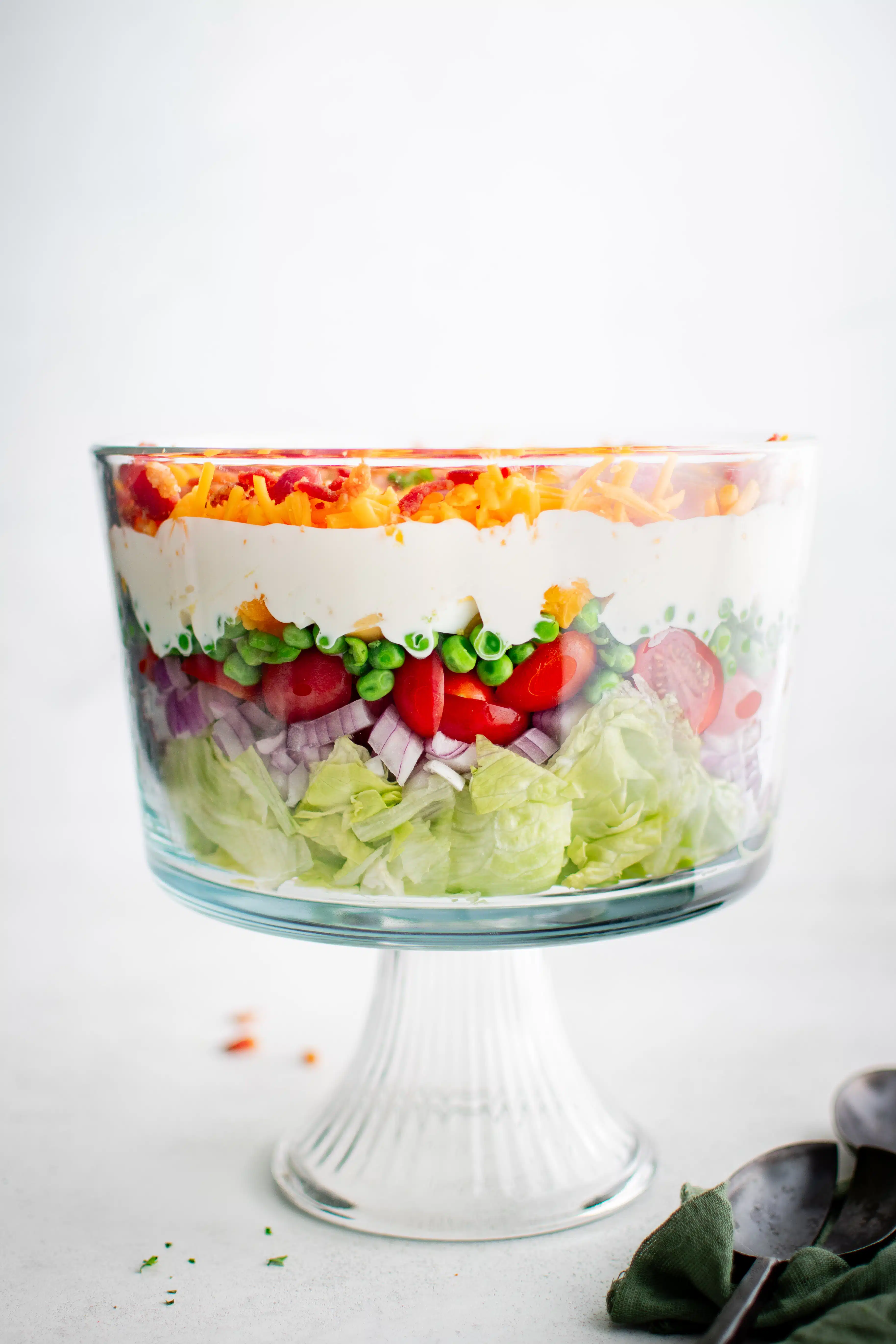 Beautifully layered traditional 7 layer salad starting with chopped iceberg lettuce on the bottom, followed by chopped red onion, sliced cherry tomatoes, green peas, chopped hard boiled eggs, creamy mayonnaise dressing, shredded cheddar cheese, and bacon bits on the top.