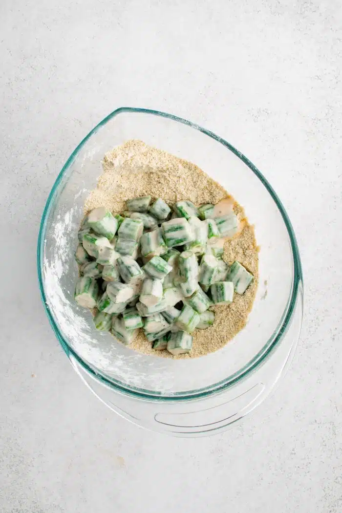 Pieces of okra coated in buttermilk added to a mixing bowl filled with all-purpose flour, cornmeal, garlic powder, onion powder, salt, pepper, and paprika.
