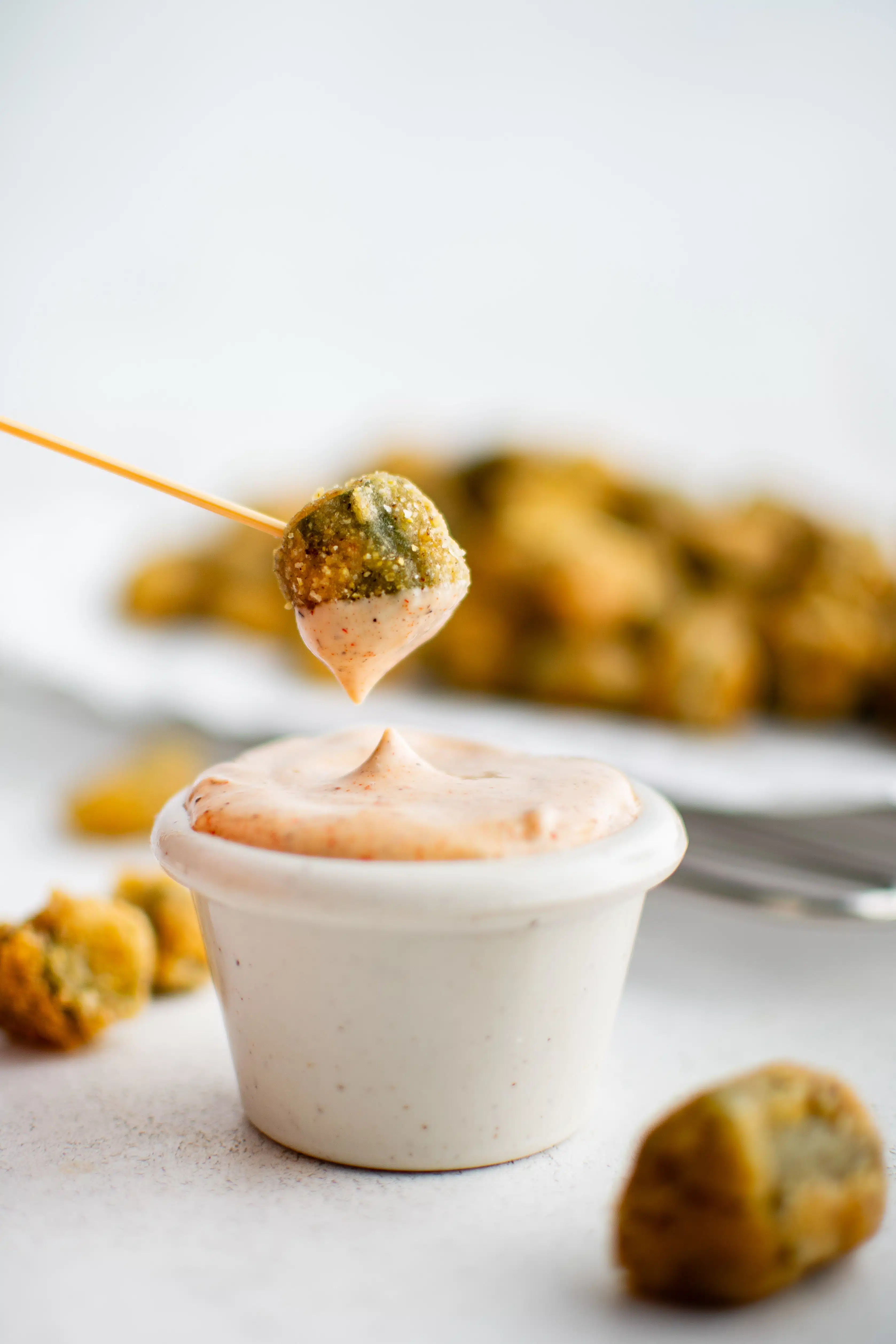 A toothpick with one piece of fried okra dipped into a small ramekin filled with creamy mayo dipping sauce.