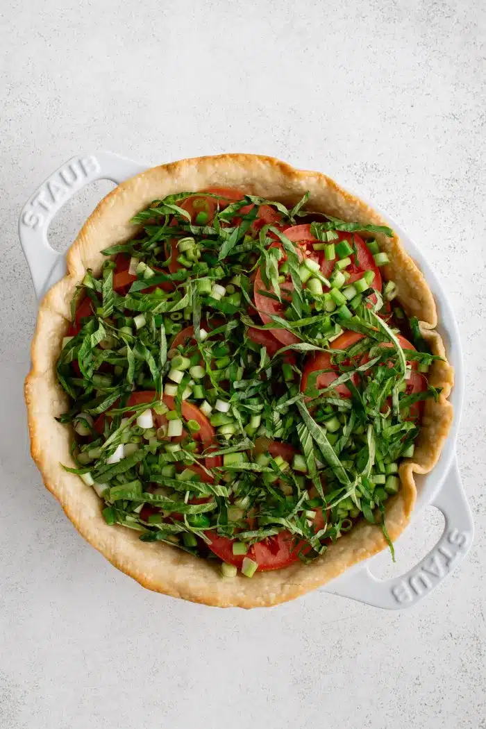 Par-baked pie crust in a white baking dish topped with freshly sliced tomatoes, basil, and green onions.
