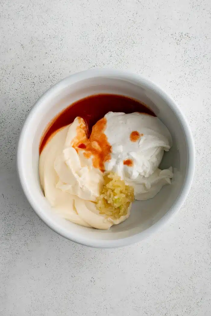 White mixing bowl filled with mayonnaise, sour cream, garlic, Frank’s hot sauce, salt and pepper.