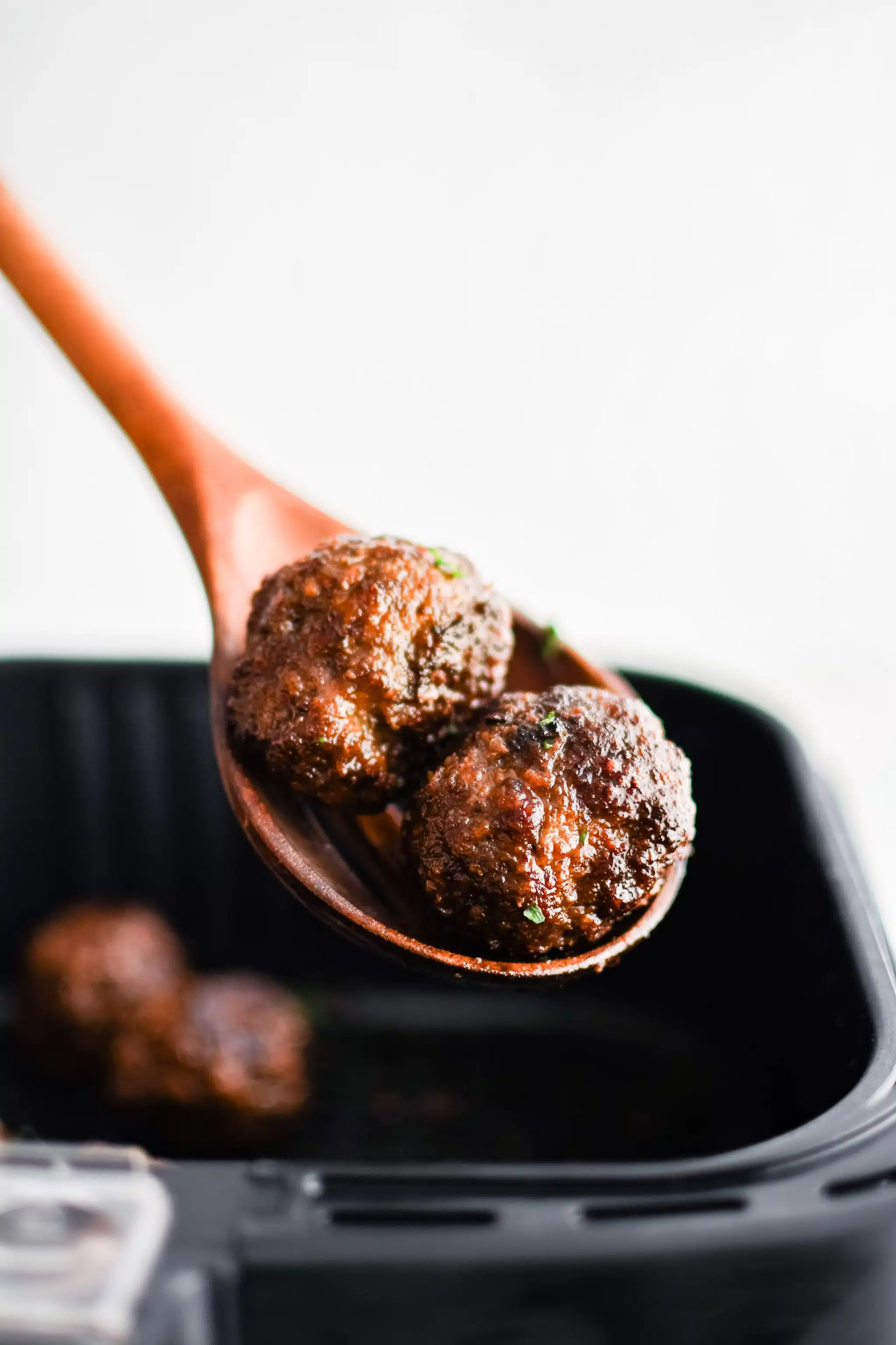 Wooden spoon holding two beef meatballs hovering above a large airfryer basket filled with the remaining cooked meatballs.