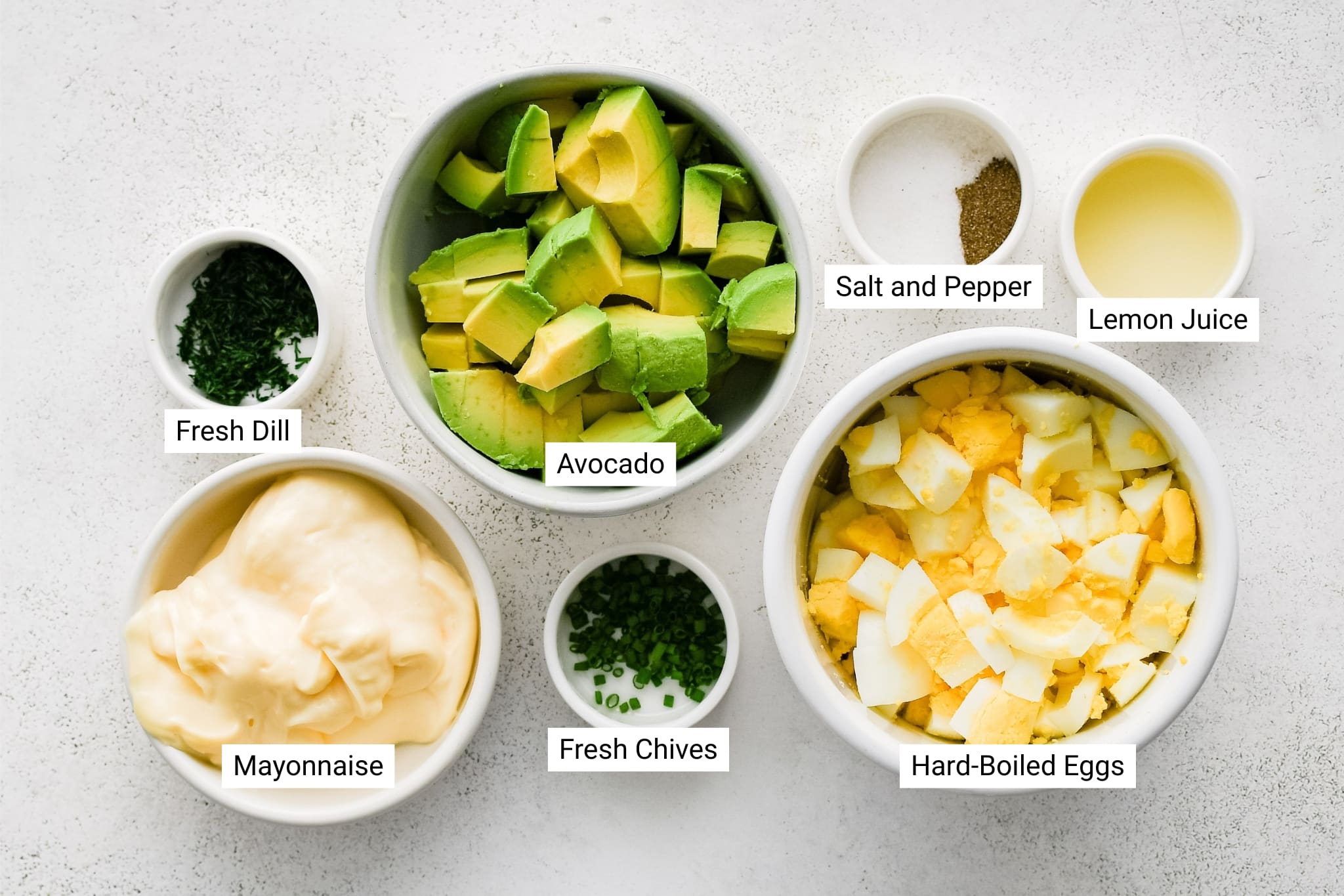 This overhead photo features the key ingredients for making avocado egg salad, each labeled clearly. Bowls of cubed avocado, chopped hard-boiled eggs, creamy mayonnaise, freshly chopped dill and chives, lemon juice, and salt and pepper are arranged on a white background.