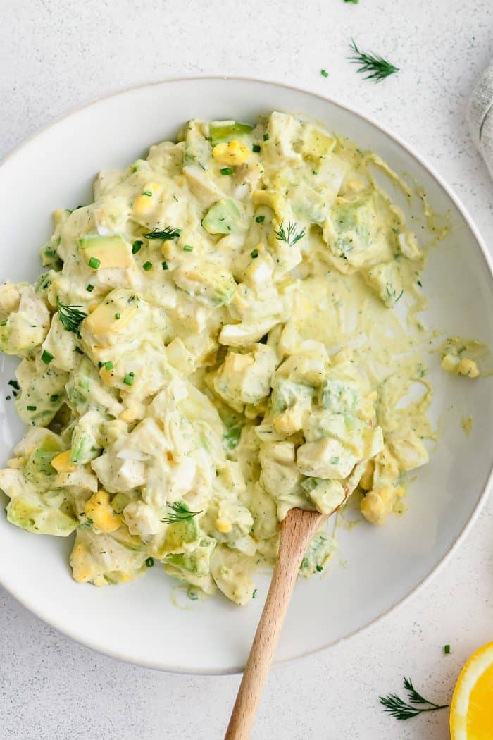 A close-up, overhead shot of creamy avocado egg salad in a white bowl. The salad features chunky pieces of avocado and hard-boiled egg, mixed with a rich, herby dressing, and garnished with fresh dill and chives. A wooden spoon rests in the salad.