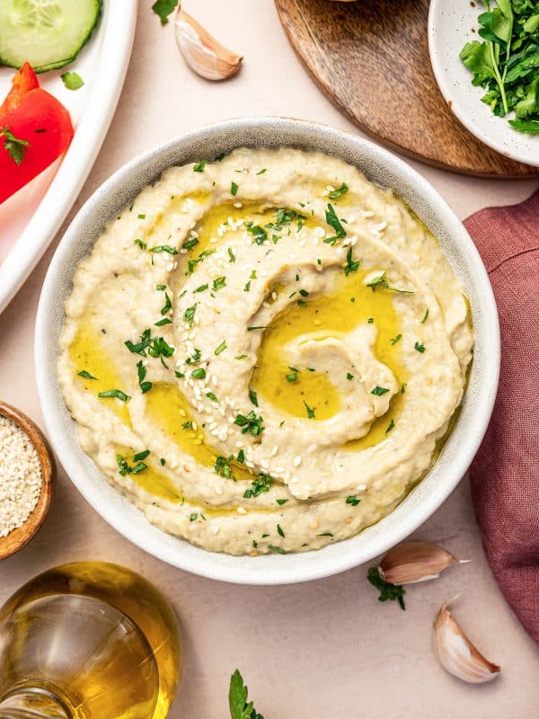 A bowl of smooth baba ganoush is swirled with extra virgin olive oil and garnished with freshly chopped parsley and sesame seeds. Fresh cucumber, garlic cloves, and parsley surround the bowl.