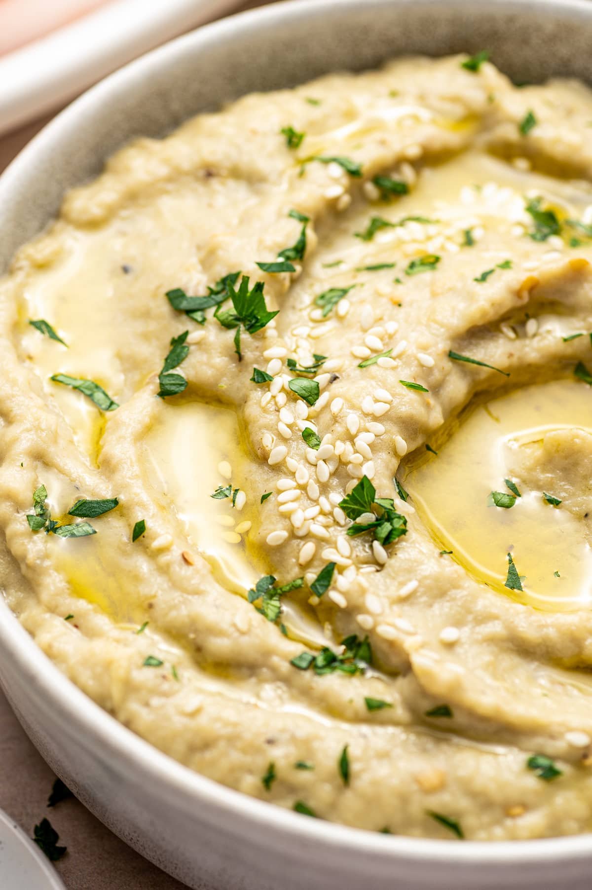 An extreme closeup shot of creamy baba ganoush focuses on the silky-smooth texture, highlighting the glistening olive oil, white sesame seeds, and flecks of green parsley.