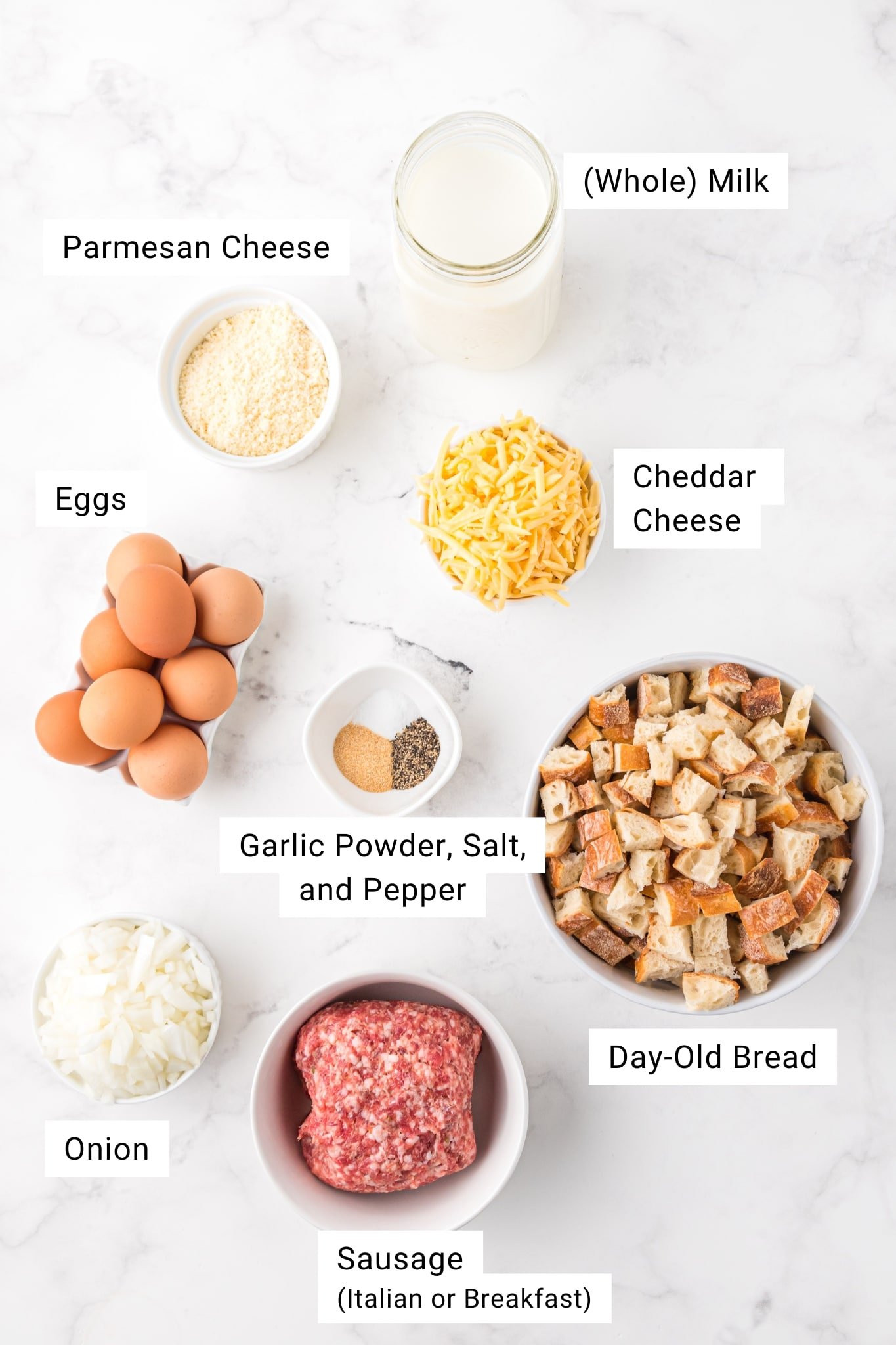 Overhead view of measured ingredients for breakfast casserole including cubed day-old bread, sausage, eggs, milk, onions, parmesan, cheddar cheese, salt, pepper, and garlic powder.