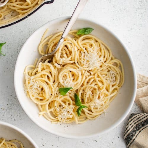 White shallow pasta bowl filled with cacio e pepe garnished with grated parmesan cheese, freshly cracked black pepper, and four fresh basil leaves.