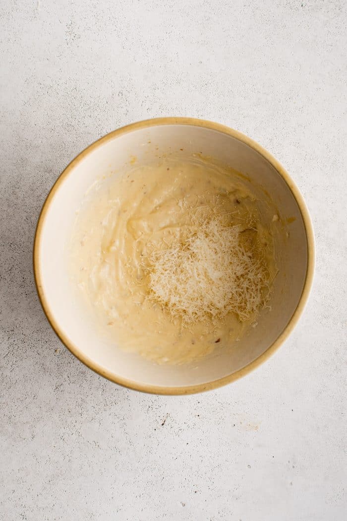 Shredded parmesan cheese added to a small bowl filled with creamy mixture of egg yolk, mayonnaise, minced anchovies, garlic, lemon juice, mustard, and salt.
