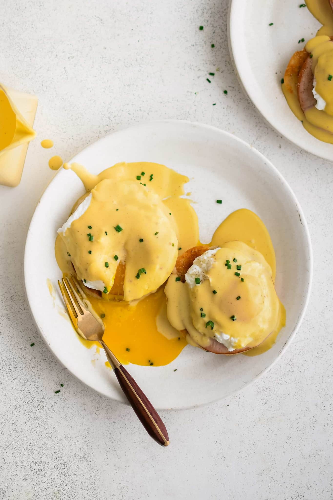 Two Eggs Benedict with split and drippy poached egg, Canadian bacon, and hollandaise sauce on an English muffin on a white plate.