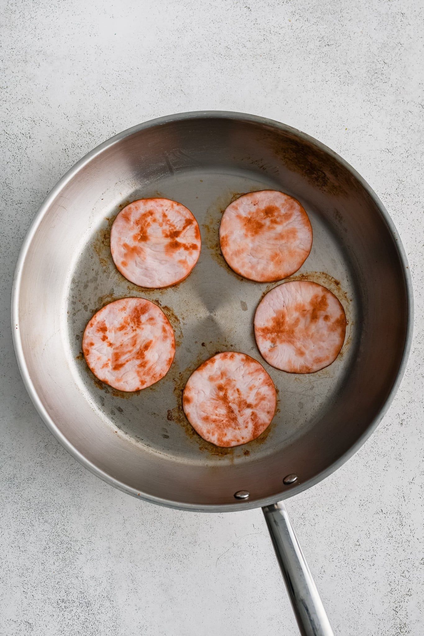 Five slices of Canadian bacon cooking in a large skillet.