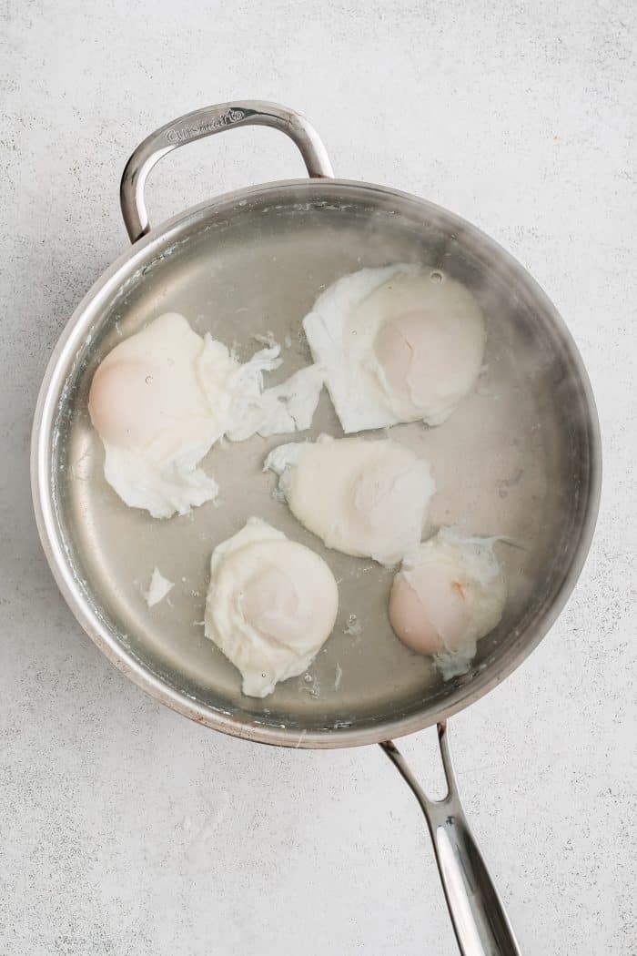 FIve eggs poaching in a large wide pan filled with simmering water.