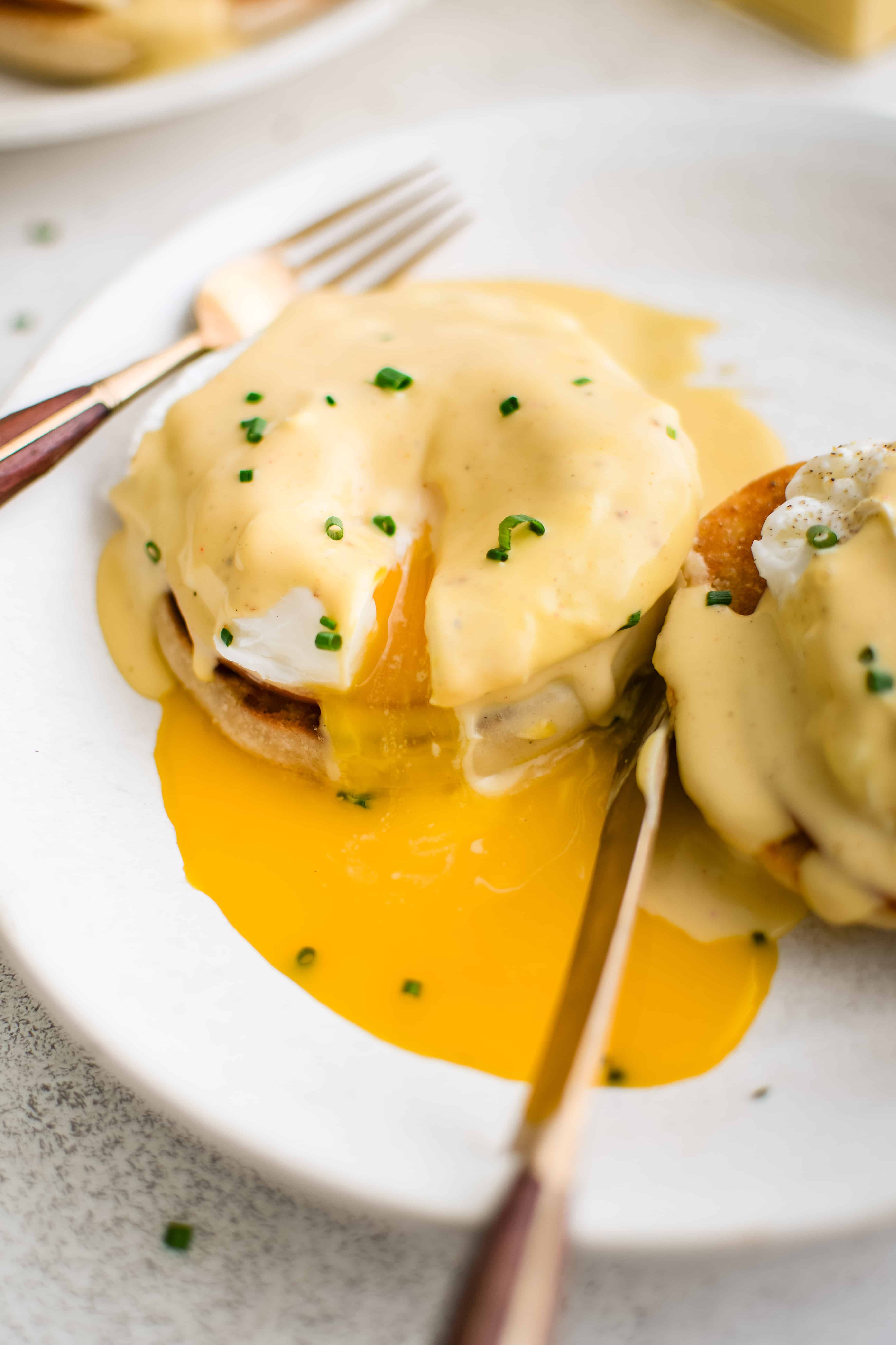 Eggs Benedict with split and drippy poached egg, Canadian bacon, and hollandaise sauce on an English muffin on a white plate.