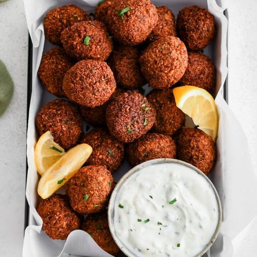 Platter lined with parchment paper and filled with golden brown fried falafel balls with a side of tzatziki and lemon wedges.