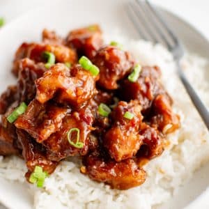 A close-up view of General Tso's Chicken served on a pristine white plate, highlighting the juicy, crispy chicken chunks drenched in a rich, caramelized sauce, accompanied by a side of steamed white rice, the dish garnished with delicate green onion slivers.