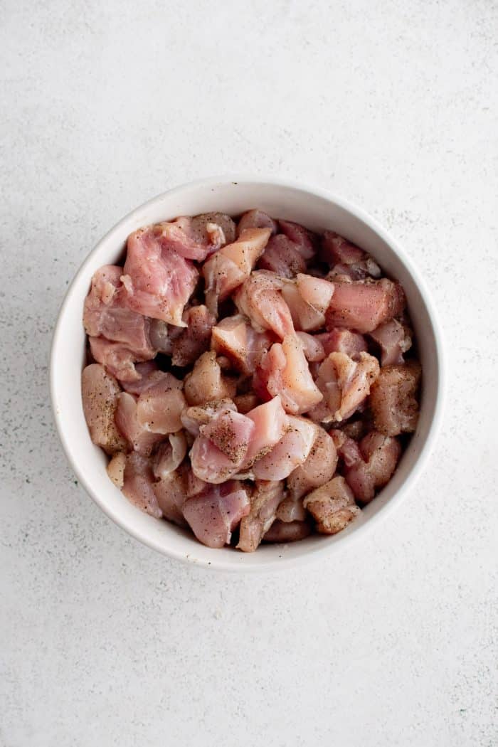 Large white mixing bowl filled with chunks of chicken thighs seasoned with salt and black pepper.