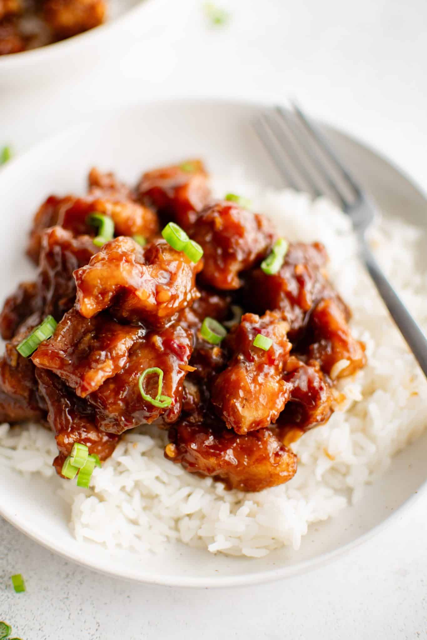 A close-up view of General Tso's Chicken served on a pristine white plate, highlighting the juicy, crispy chicken chunks drenched in a rich, caramelized sauce, accompanied by a side of steamed white rice, the dish garnished with delicate green onion slivers.