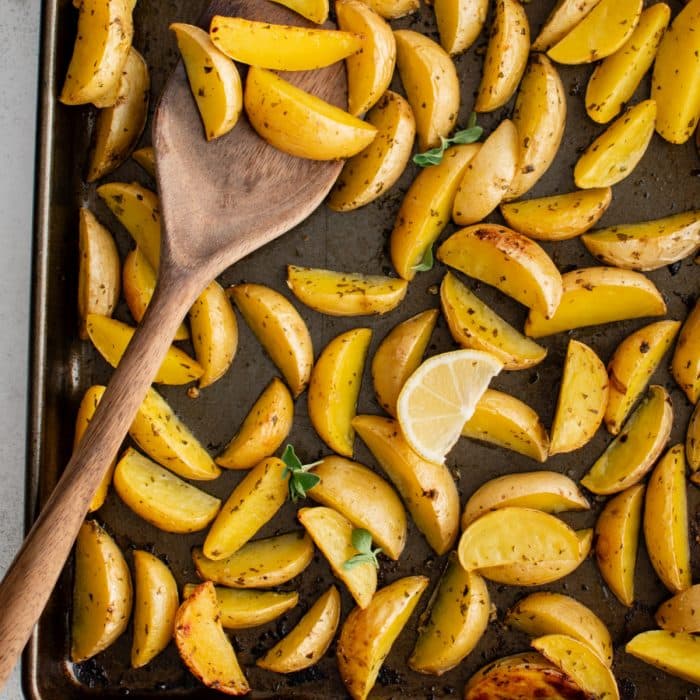 Golden roasted Greek potatoes seasoned with fresh parsley and oregano, lemon juice, and garlic, on a baking sheet with lemon wedges.